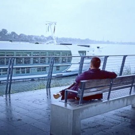 River Crew Mitglied sitzt auf einer Bank am Ufer eines Flusses mit Blick auf ein Flusskreuzfahrtschiff von Phoenix Reisen