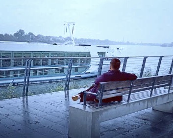 River Crew Mitglied sitzt auf einer Bank am Ufer eines Flusses mit Blick auf ein Flusskreuzfahrtschiff von Phoenix Reisen
