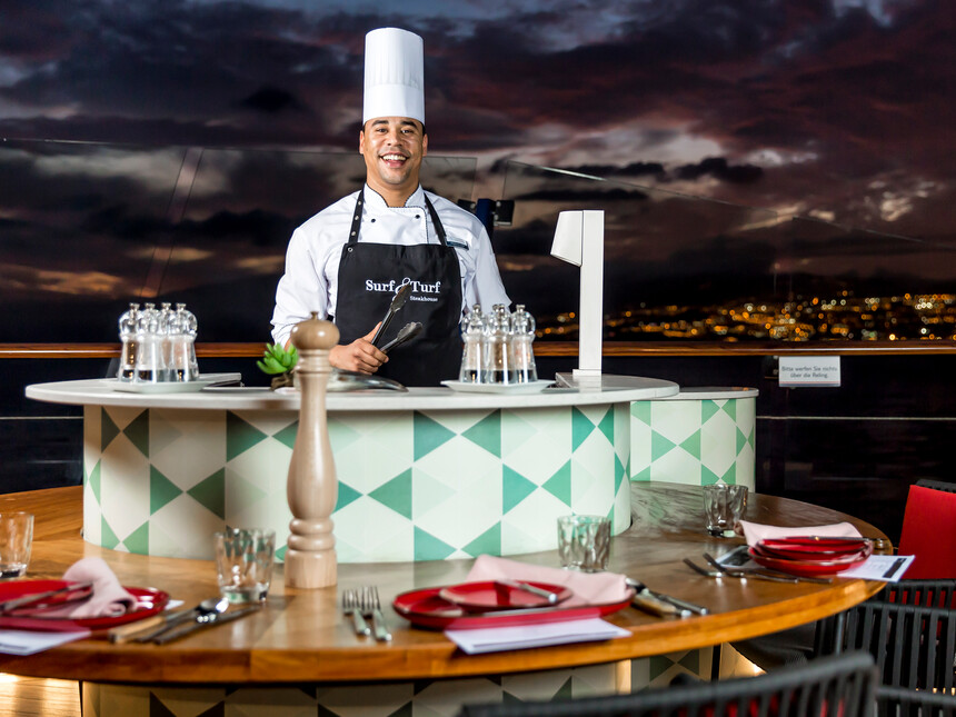 Koch auf Mein Schiff macht live cooking an der Reling mit Blick auf eine Insel bei Nacht