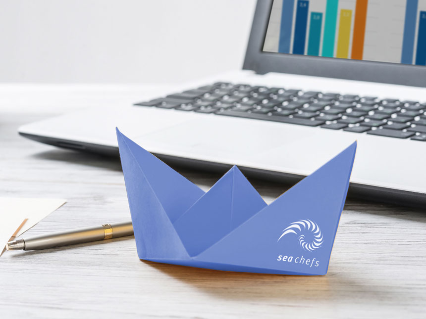 Papierschiffchen in blau mit sea chefs Logo vor einem Laptop auf einem Schreibtisch